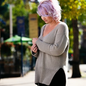 May include: A woman with lavender hair wearing a light brown, long-sleeved, knitted sweater with a v-neck and a side slit. She is wearing black pants.