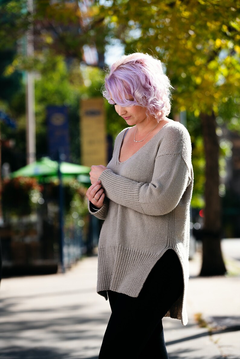 May include: A woman with lavender hair wearing a light brown, long-sleeved, knitted sweater with a v-neck and a side slit. She is wearing black pants.
