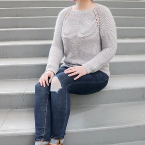 May include: A woman wearing a light gray knitted sweater with a delicate lace pattern, blue jeans with a distressed knee, and brown ankle boots. She is sitting on a set of concrete stairs.