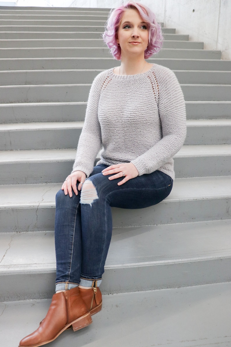 May include: A woman wearing a light gray knitted sweater with a delicate lace pattern, blue jeans with a distressed knee, and brown ankle boots. She is sitting on a set of concrete stairs.