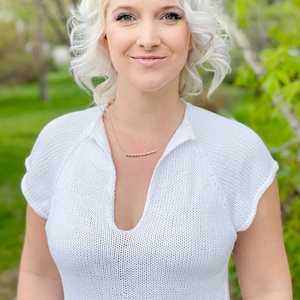 May include: A woman with blonde hair is wearing a white, knitted, short-sleeved top with a deep V-neck. She is wearing a silver necklace with small beads.
