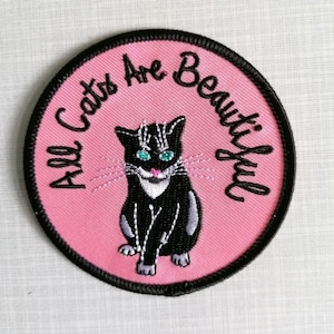 Könnte beinhalten: Ein rosa und schwarzer gestickter Aufnäher mit einer schwarzen Katze mit weißen Markierungen und grünen Augen. Der Text "All Cats Are Beautiful" ist in schwarzer kursivschrift um die Katze gestickt.