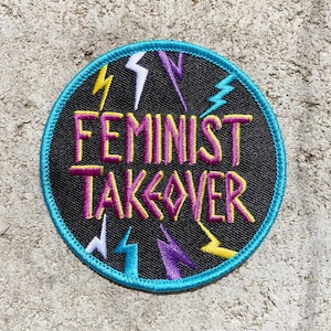 Peut inclure: Écusson rond avec l'inscription "FEMINIST TAKEOVER" en lettres jaunes et violettes. Le fond noir est orné d'éclairs jaunes, blancs et turquoise. L'écusson est bordé d'une couture turquoise.