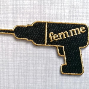 Könnte beinhalten: Schwarzer und goldener gestickter Aufnäher einer Bohrmaschine mit dem Wort "femme" darauf.