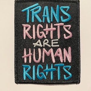 Op de afbeelding: Een zwarte vierkante patch met geborduurde tekst. De woorden "TRANS RIGHTS" zijn in blauw, "ARE" in wit en "HUMAN RIGHTS" in roze en blauw. De patch heeft een zwarte rand.