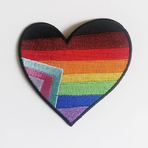 Op de afbeelding: Een regenboogvormige, hartvormige geborduurde patch met een zwarte omtrek. De regenboogkleuren zijn rood, oranje, geel, groen, blauw en paars. De patch is gemaakt van stof en heeft een textuur.