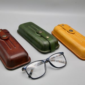 May include: Three leather eyeglass cases in brown, green, and yellow, with a pair of blue-framed glasses in the foreground.