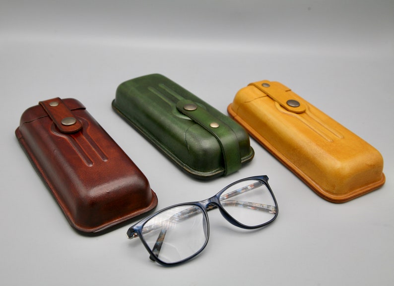 May include: Three leather eyeglass cases in brown, green, and yellow, with a pair of blue-framed glasses in the foreground.