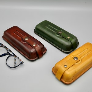 May include: Three leather eyeglass cases in brown, green, and yellow. Each case has a flap with a button closure and a decorative design.