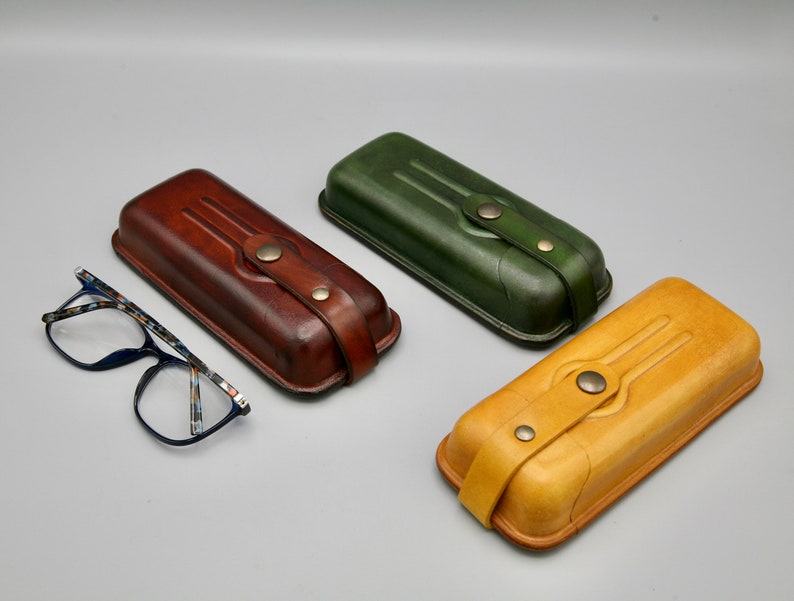 May include: Three leather eyeglass cases in brown, green, and yellow. Each case has a flap with a button closure and a decorative design.