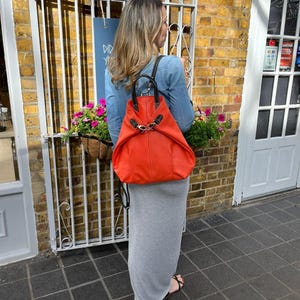 May include: A vibrant orange leather backpack with black straps and silver clasps. The bag is carried by a person wearing a denim shirt and a grey maxi skirt. The background features a brick wall, a white metal gate, and a sign.