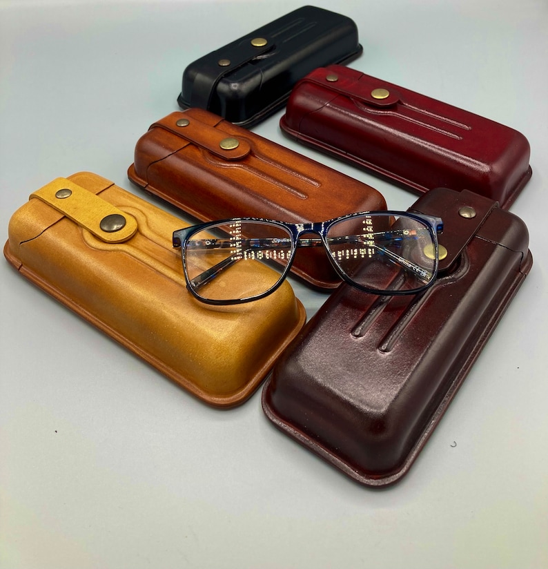 May include: Five leather eyeglass cases in different colors, including brown, tan, black, and burgundy. A pair of blue-framed eyeglasses is resting on top of two of the cases.