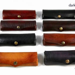 Puede incluir: Diez estuches para bolígrafos de cuero en diferentes colores, incluyendo naranja oscuro, rojo oscuro, negro, amarillo, verde oscuro, marrón oscuro, coñac y morado oscuro.