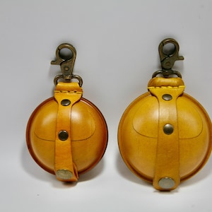 May include: Two round, yellow leather pouches with brass-coloured clasps and hardware. Each pouch has a strap and a snap closure. The pouches are similar in size, with one slightly smaller than the other.