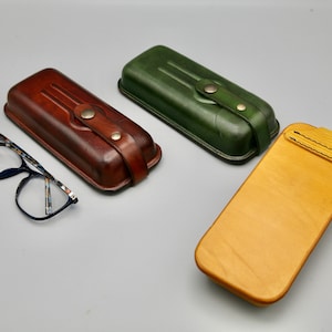 May include: Three different styles of leather eyeglass cases. One is brown, one is green, and one is wood with a yellow stain.