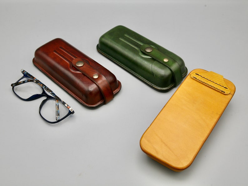 May include: Three different styles of leather eyeglass cases. One is brown, one is green, and one is wood with a yellow stain.