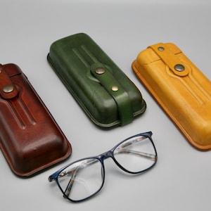 May include: Three leather eyeglass cases in brown, green, and tan. The cases have a flap closure with a button. A pair of blue-framed eyeglasses are in the center of the image.