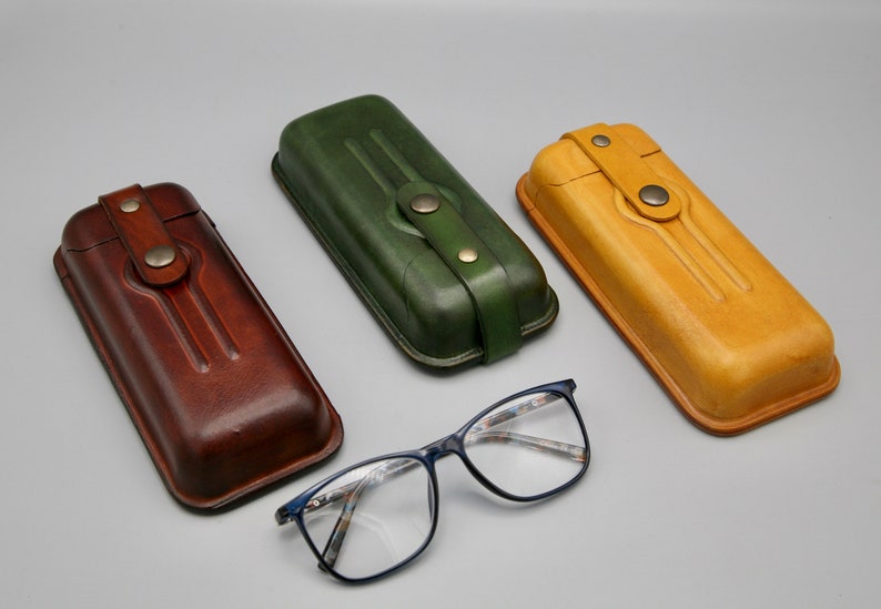 May include: Three leather eyeglass cases in brown, green, and tan. The cases have a flap closure with a button. A pair of blue-framed eyeglasses are in the center of the image.