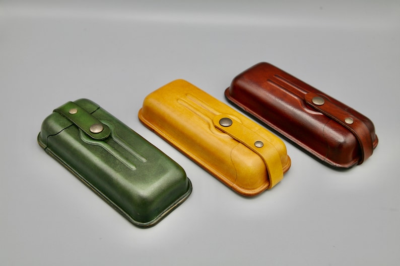 May include: Three leather cases in green, yellow, and brown. Each case has a flap with a strap and a metal button closure.