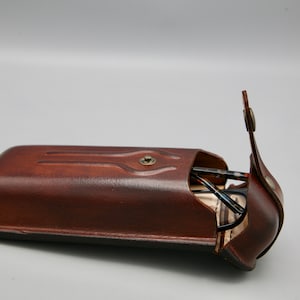 May include: A brown leather glasses case with a flap that opens to reveal a pair of glasses inside.