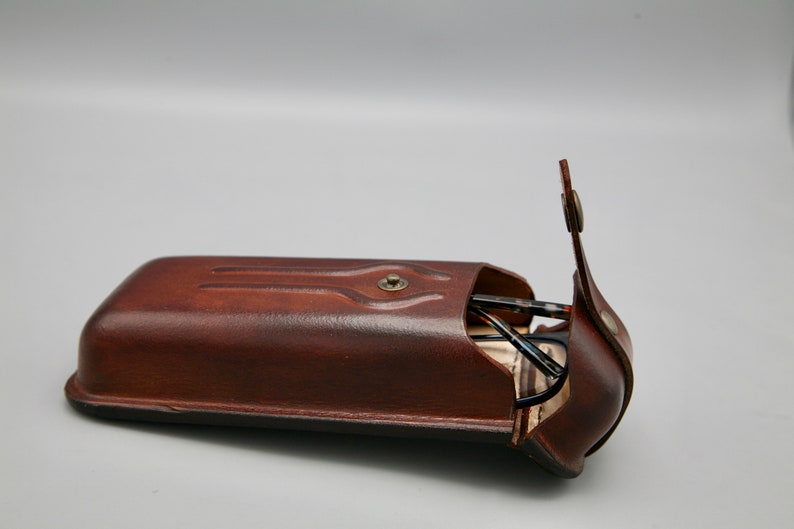 May include: A brown leather glasses case with a flap that opens to reveal a pair of glasses inside.