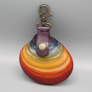 May include: A colorful, hand-painted leather keychain pouch. The pouch is shaped like a clam shell and features a rainbow of colors, including red, orange, yellow, blue, and purple. It has a purple leather strap and a bronze-colored clasp.