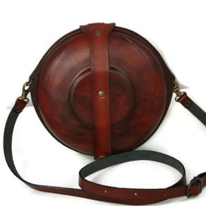 May include: A brown leather round crossbody bag with a long adjustable strap. The bag has a flap closure and a decorative strap across the front.