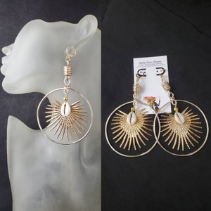 May include: Gold and silver hoop earrings with a white cowrie shell and gold accents. The earrings are handmade and feature a sunburst design.