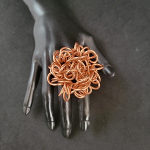 May include: A large, copper-colored ring made of twisted wire. The ring is displayed on a black mannequin hand.