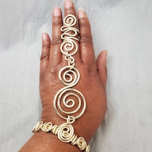 Puede incluir: Una pulsera de mano de alambre de color dorado con un anillo para el dedo conectado a una pulsera mediante una serie de diseños en espiral. La pulsera y el anillo están conectados por una cadena de alambre en espiral. La joya se muestra en una mano.