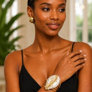 May include: A gold-toned cuff bracelet featuring a central white cowrie shell with dark brown spots, wrapped in gold wire. The bracelet is worn with matching gold-toned dome-shaped stud earrings. The jewelry is paired with a black spaghetti strap top.