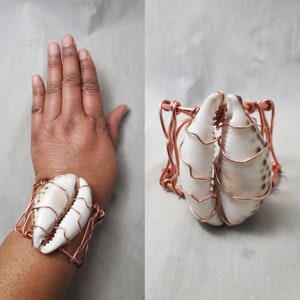 May include: A copper wire bracelet with two white seashells in the shape of crab claws. The bracelet is wrapped around a person's wrist.