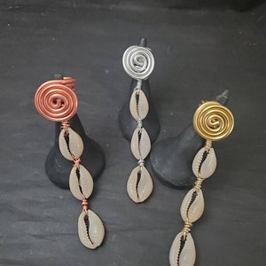 May include: Three hair jewelry pieces with a spiral design in gold, silver, and copper. Each piece has a chain of white cowrie shells hanging from the spiral.