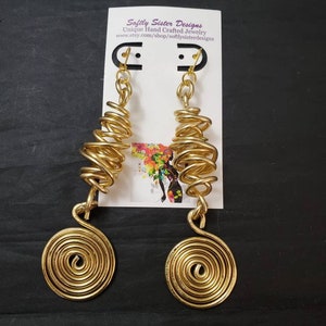 以下が含まれることがあります： 螺旋模様のゴールドトーンのイヤリング。イヤリングはワイヤー製で、フック留めです。イヤリングは、「Softly Sister Designs Unique Hand Crafted Jewelry www.etsy.com/shop/softlysisterdesigns」というテキストが書かれた白いカードに飾られています。