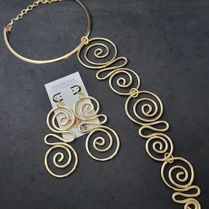 May include: A gold-colored jewelry set featuring a necklace and earrings. The necklace has a choker-style base with a chain clasp and a decorative spiral design. The earrings also have a spiral design. The jewelry is displayed on a dark background.