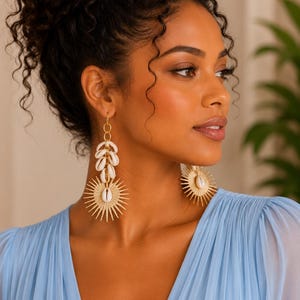 May include: Gold-toned drop earrings featuring a vertical arrangement of small white cowrie shells suspended above a larger, circular sunburst-shaped pendant with a central cowrie shell. The earrings have a metallic finish and a dangling design.