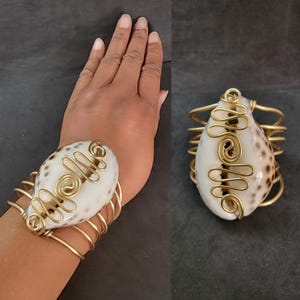 Puede incluir: Un brazalete de alambre dorado envuelto alrededor de una concha de mar blanca y marrón. El alambre tiene la forma de la palabra "hola".