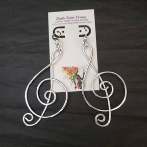 May include: Silver wire earrings shaped like treble clefs. The earrings are hanging from a white card with the text "Softly Sister Designs Unique Hand Crafted Jewelry www.etsy.com/shop/softlysisterdesigns".
