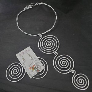 May include: A silver-coloured jewellery set comprising a necklace, earrings, and a bracelet. The necklace and earrings feature a spiral design. The bracelet has a twisted wire design. The jewellery set is displayed on a black surface.
