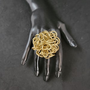 May include: A gold-colored ring made of twisted wire, resembling a tangled ball of yarn. The ring is worn on a black mannequin hand.