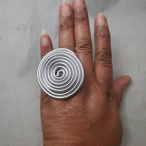 May include: A large silver ring with a spiral design. The ring is worn on the index finger of a hand.