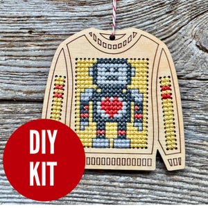 May include: Wooden DIY kit in the shape of a sweater with a cross-stitched robot design. The robot has a red heart and is surrounded by yellow, gray, and black cross-stitch. The kit includes the text "DIY KIT" on a red circle.