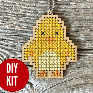 May include: DIY cross stitch kit featuring a yellow chick with orange feet. The chick is on a wooden background.