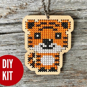 May include: DIY kit for a cross-stitch ornament featuring a cute orange, black, and white tiger. The ornament is made of wood and comes with a string for hanging.