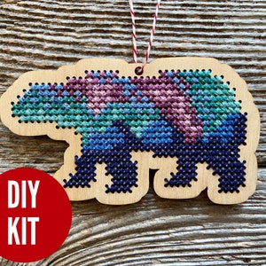 May include: DIY cross-stitch kit featuring a wooden bear ornament with a colorful pixelated design. The bear is outlined in dark blue and has a pink, blue, and green color scheme.