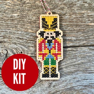 May include: Wooden ornament in the shape of a nutcracker, featuring a cross-stitch design. The ornament has a yellow, red, blue, green, and black color scheme. The text "DIY KIT" is on a red circle in the lower left corner.
