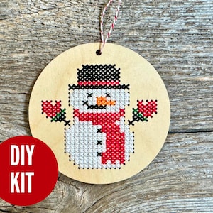 May include: A round wooden ornament with a cross-stitched snowman design. The snowman has a black hat, red scarf, and holds red floral accents. The ornament has a red and white string for hanging and a red circle with "DIY KIT" text.