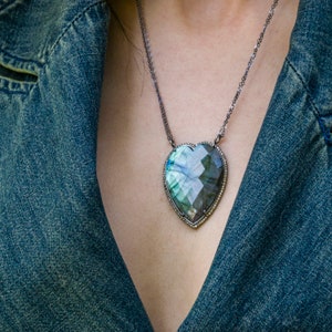 Puede incluir: Un collar de cadena de plata con un gran colgante en forma de coraz&oacute;n. El colgante est&aacute; hecho de una piedra preciosa verde oscuro y gris con un dise&ntilde;o facetado y un borde plateado.