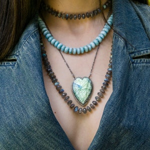 Puede incluir: Un collar de capas con un colgante en forma de coraz&oacute;n con una piedra preciosa verde y blanca. El collar est&aacute; hecho de plata y presenta una variedad de cuentas, incluyendo azules, grises y negras.