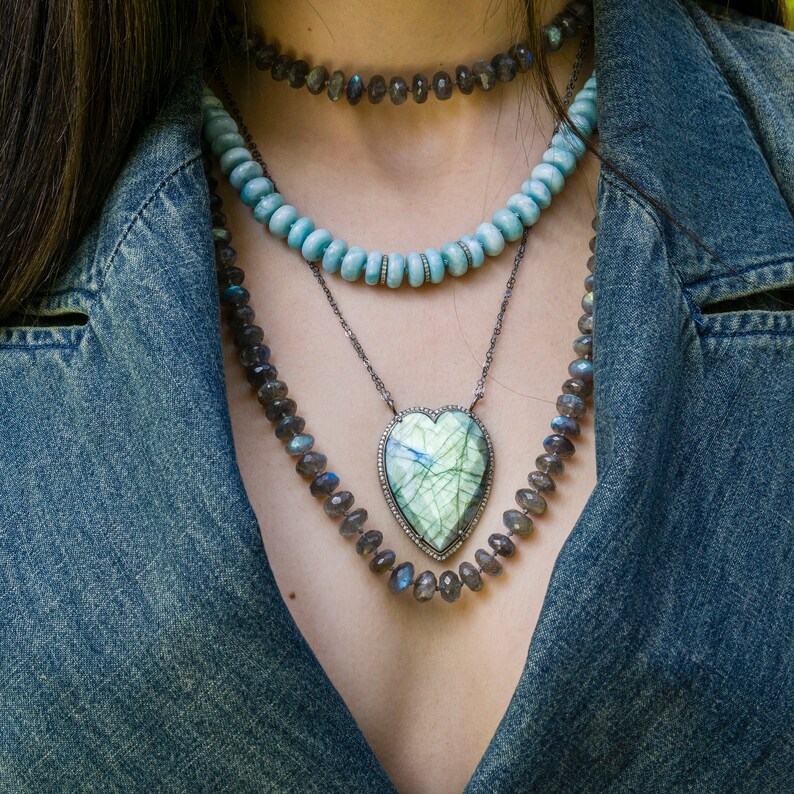 Puede incluir: Un collar de capas con un colgante en forma de coraz&oacute;n con una piedra preciosa verde y blanca. El collar est&aacute; hecho de plata y presenta una variedad de cuentas, incluyendo azules, grises y negras.